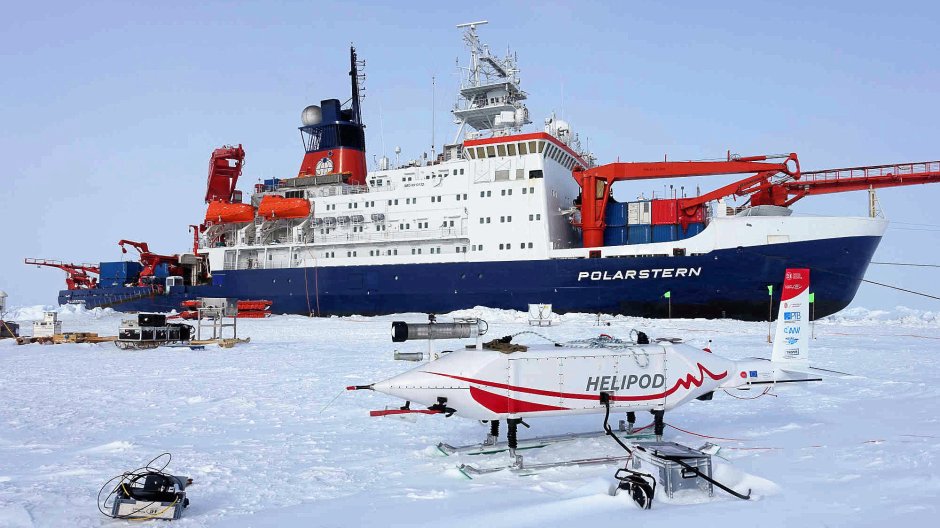 The photo shows the helicopter-borne sonde HELiPOD standing on the snow-covered sea ice. The research vessel Polarstern is in the background. Photo: Falk Pätzold / TU Braunschweig