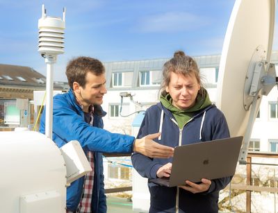 Two young men stand on the roof of the institute behind the micro rain radar and look on a laptop. Photo: Swen Reichhold 