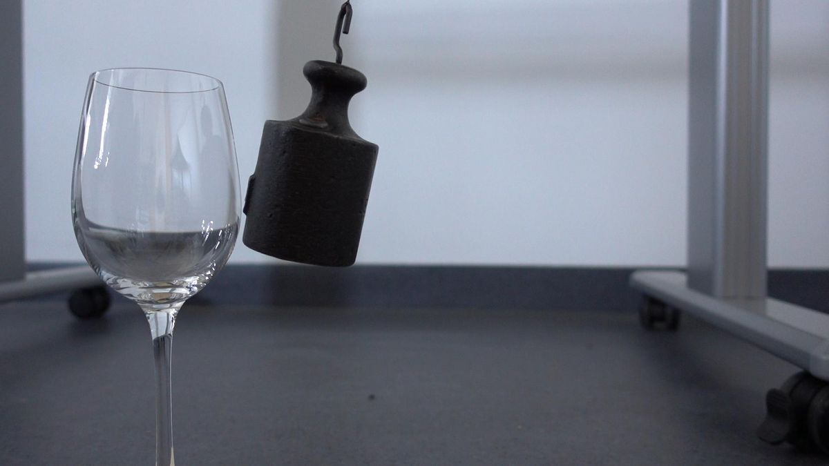 Ausschnitt aus einem Experiment mit Massestück an Faden und Weinglas. Das 1 kg Massestück berührt in einer Pendelbewegung fast das Weinglas. Foto: Kilian Peisker
