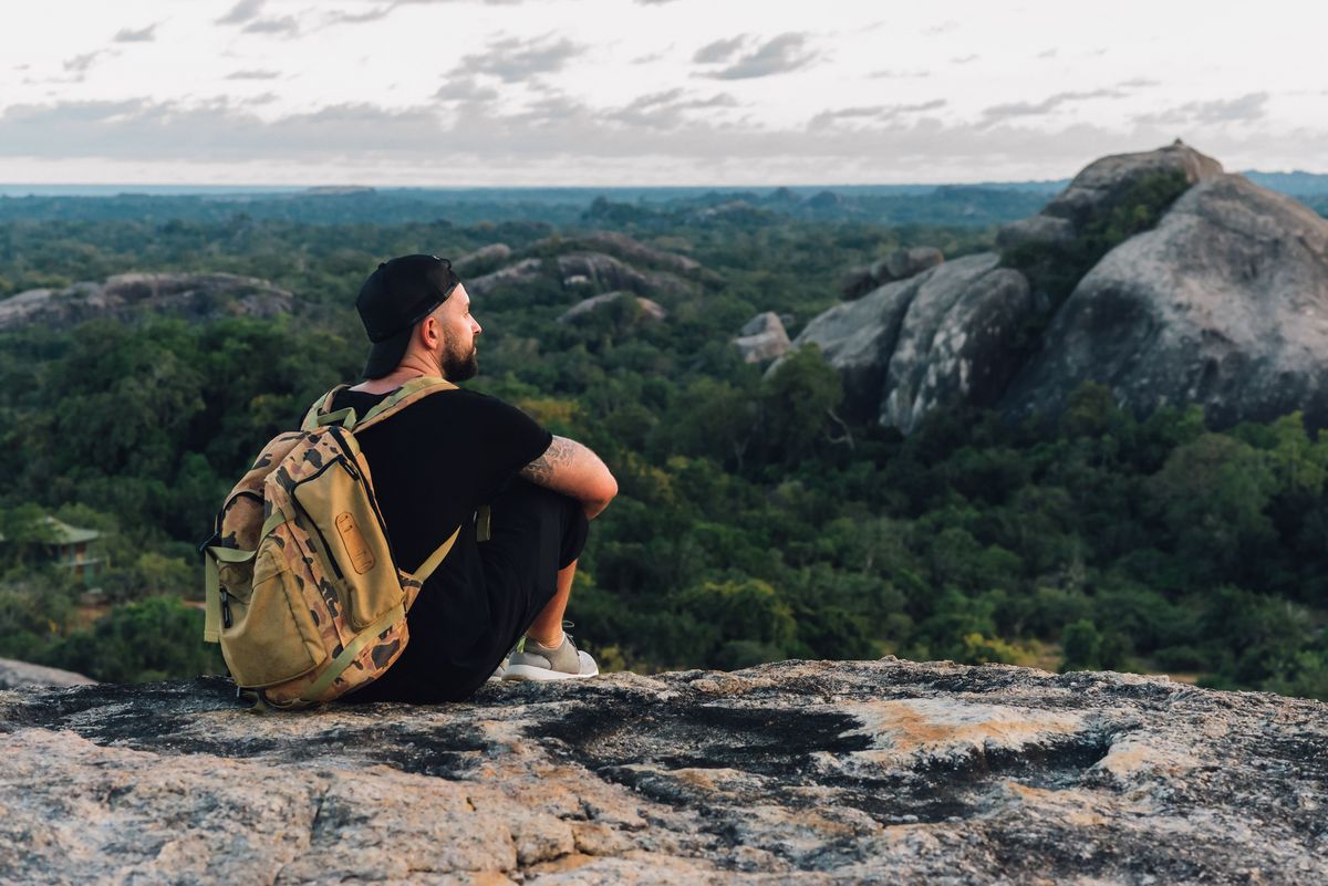 zur Vergrößerungsansicht des Bildes: Ein junger Mann sitzt auf einem Stein und guckt in die Ferne auf eine Berglandschaft.