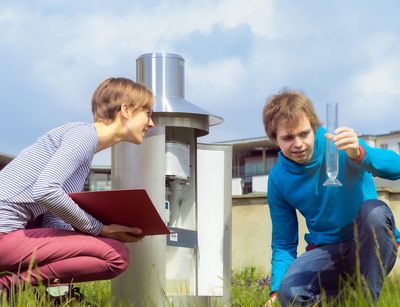Zwei Studierden sitzen auf der Wetterwiese. Ein Studierender hät einen Glaszylinder hoch und eine Studierende liest ab. Foto. Swen Reichhold