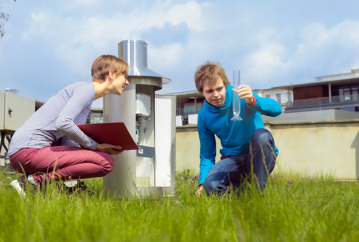 Two students are sitting on the weather meadow. A student holds up a glass cylinder and a student takes a reading. Photo. Swen Reichhold