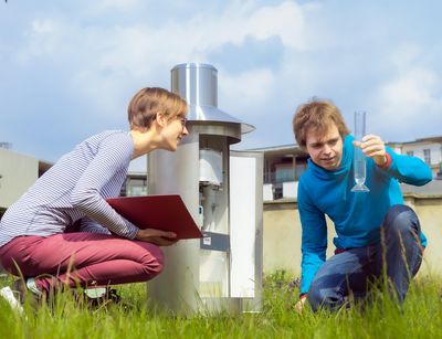 Two students are sitting on the weather meadow. A student holds up a glass cylinder and a student takes a reading. Photo. Swen Reichhold