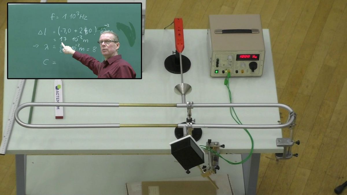 Auf einem Tisch ist ein Experiment aufgebaut mit einem Frequenzgenerator und einem verschiebbaren oval gebogenen Rohr, oben links ist ein Fenster mit Dozent Frank Stallmach an der Tafel, Videoausschnitt: Universität Leipzig