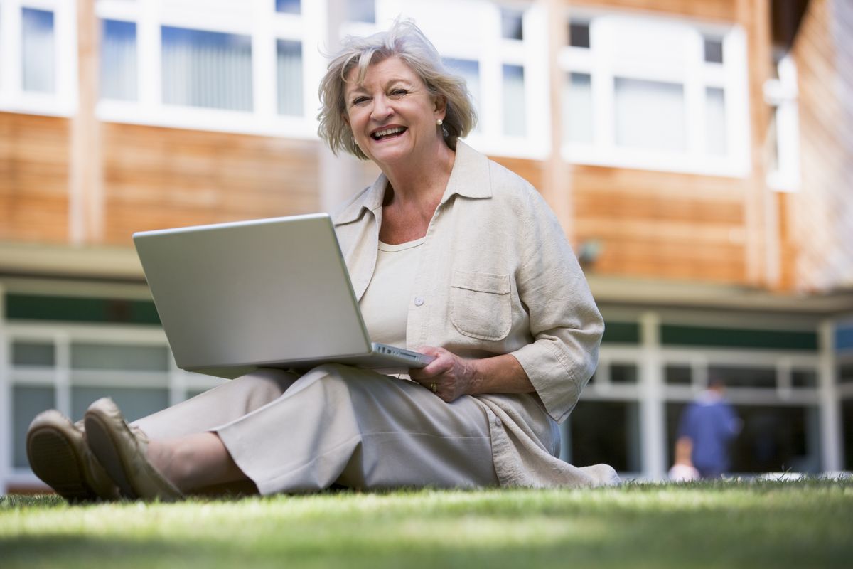zur Vergrößerungsansicht des Bildes: Eine ältere Frau sitzt mit ihrem Laptop auf einer Wiese und freut sich. Foto: Colourbox