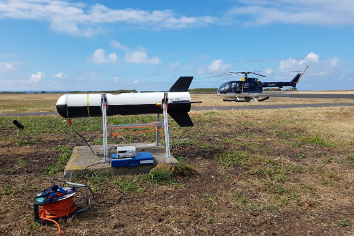 Das Bild zeigt die Messplattform SMART-HELIOS mit der Form eines Torpedos die am Boden steht und bei der Messung am Hubschrauber hängt. Foto: Felix Lauermann