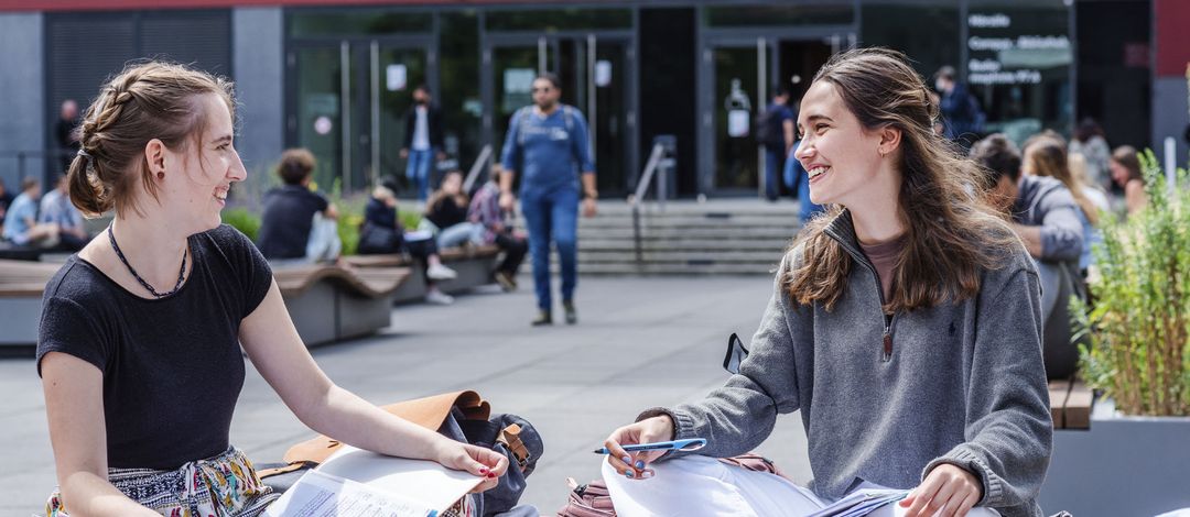 Two female students sit cross-legged on a bench in the university courtyard and talk. In the background, out of focus, people can be seen walking into the lecture hall building.